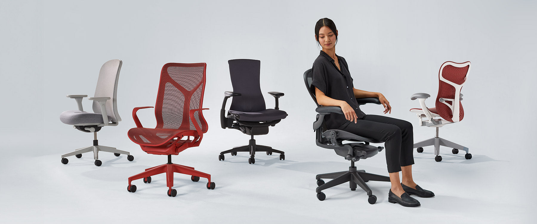 Photo des chaises Herman Miller alignées, notamment les modèles Verus blanc minéral et gris, Cosm à dossier moyen en rouge canyon, Embody noir, Aeron graphite avec une femme assise et Mirra 2 en rouge cordovan