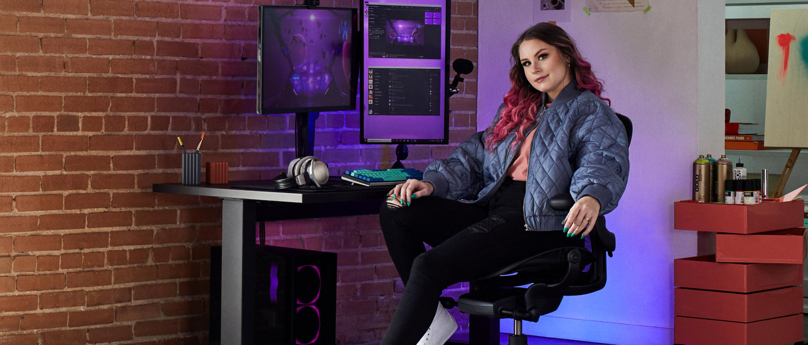 Photo de la joueuse Electra face à la caméra, assise sur une chaise de jeu Aeron noir onyx devant un bureau de jeu Nevi noir avec un bras support pour écran Ollin