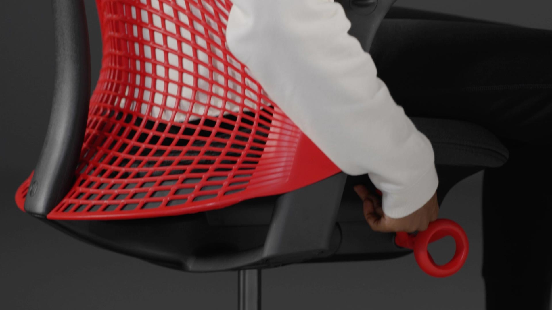 Vidéo d'une personne assise dans un fauteuil de jeu Sayl rouge et noir de Herman Miller, qui tend la main à côté du fauteuil pour régler la hauteur de l'assise.
