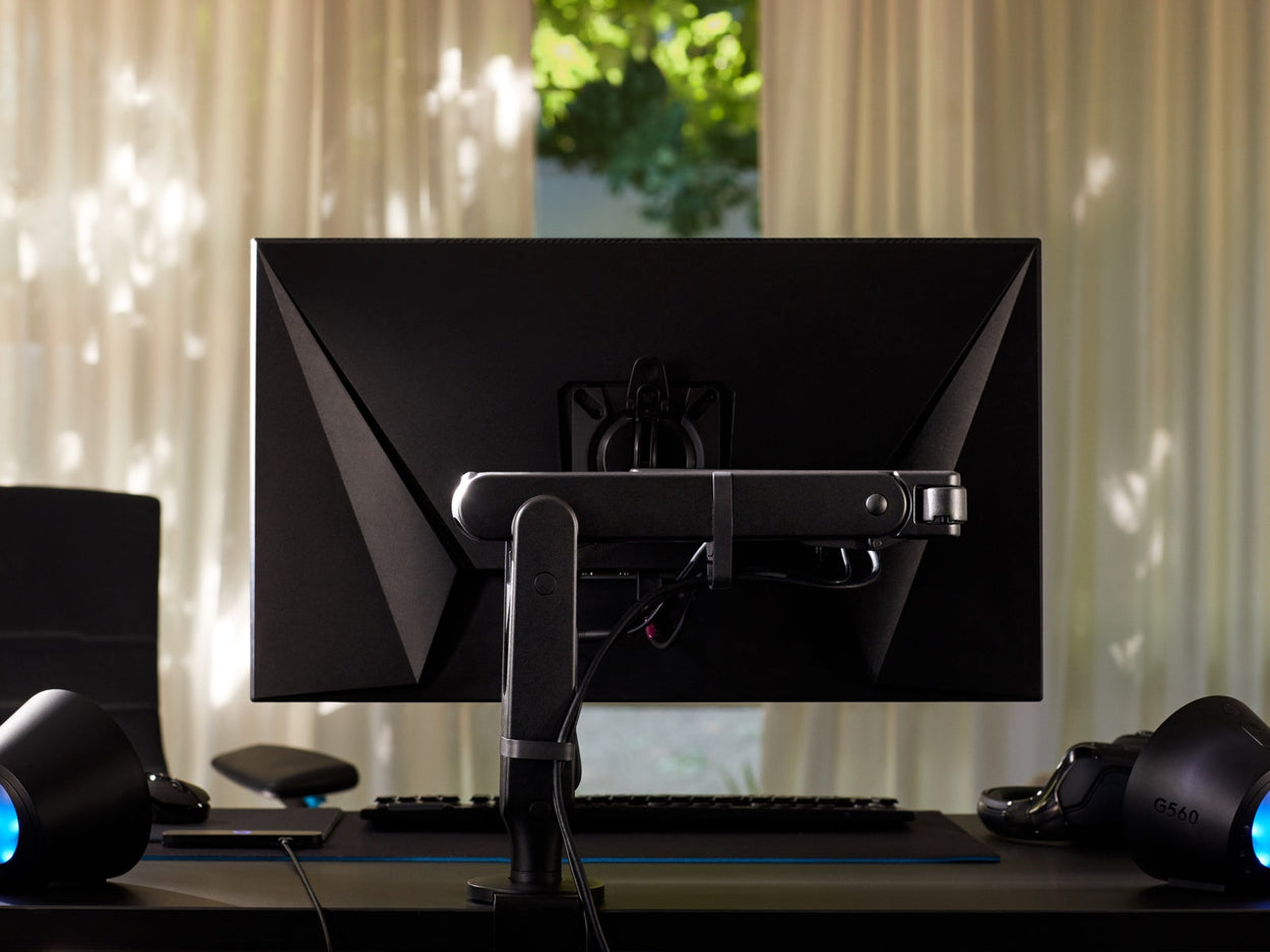 Vue arrière d'un bras de moniteur Ollin noir tenant un écran d'ordinateur et fixé à un bureau de jeu Nevi noir de Herman Miller. Installation dans une salle de jeux ou un bureau à domicile, avec des lampes, un clavier et une souris.
