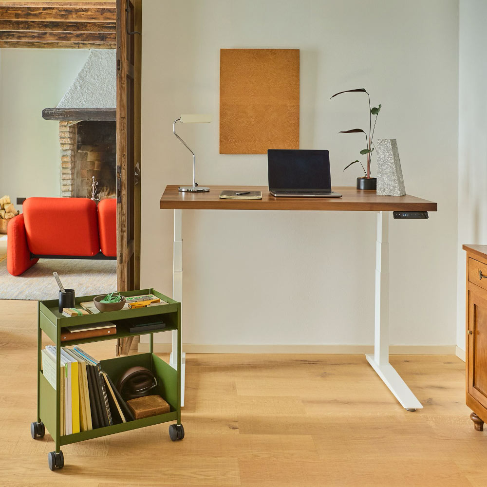 Un bureau assis-debout Herman Miller Jarvis dans sa position la plus haute, avec un chariot OE1 vert olive, un canapé chiclet Wilkes rouge et une lampe HAY Apex.
