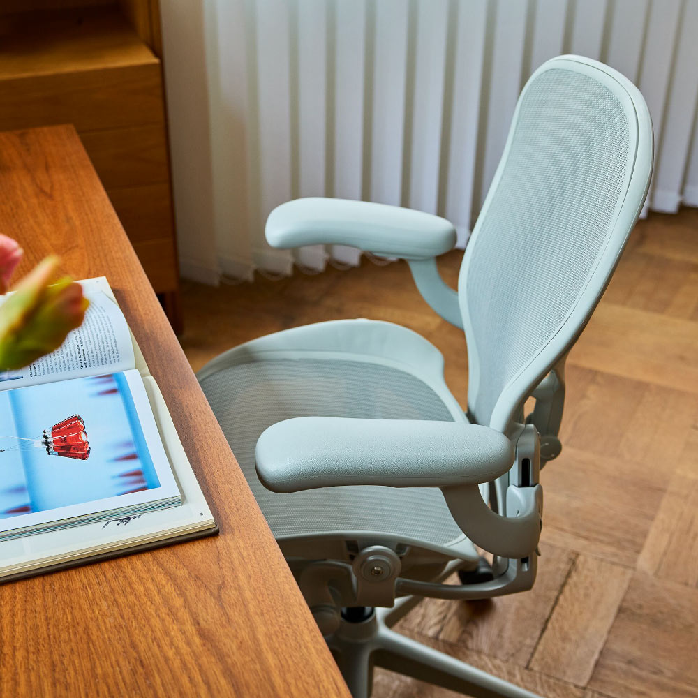 Vue rapprochée d'une chaise de bureau ergonomique Herman Miller Aeron, gris pâle et minéral, posée sur une table en bois.