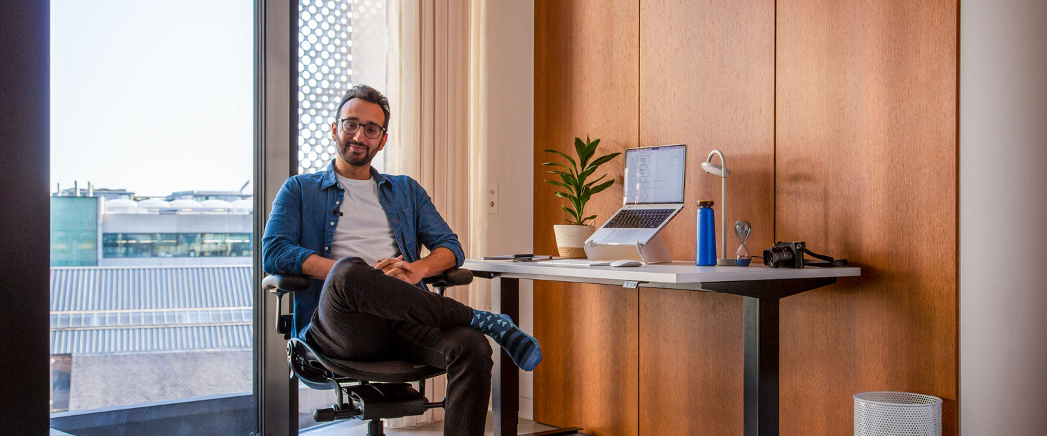 Photo d'Ali Abdaal, podcasteur, assis sur une chaise de bureau Herman Miller Aeron en graphite devant un bureau assis-debout Nevi avec des pieds noirs et un plateau blanc, un ordinateur portable posé sur un support Oripura