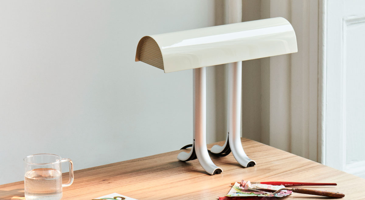 Une lampe de table Anagram crème et argentée de HAY, placée dans le coin arrière d'un bureau en bois.