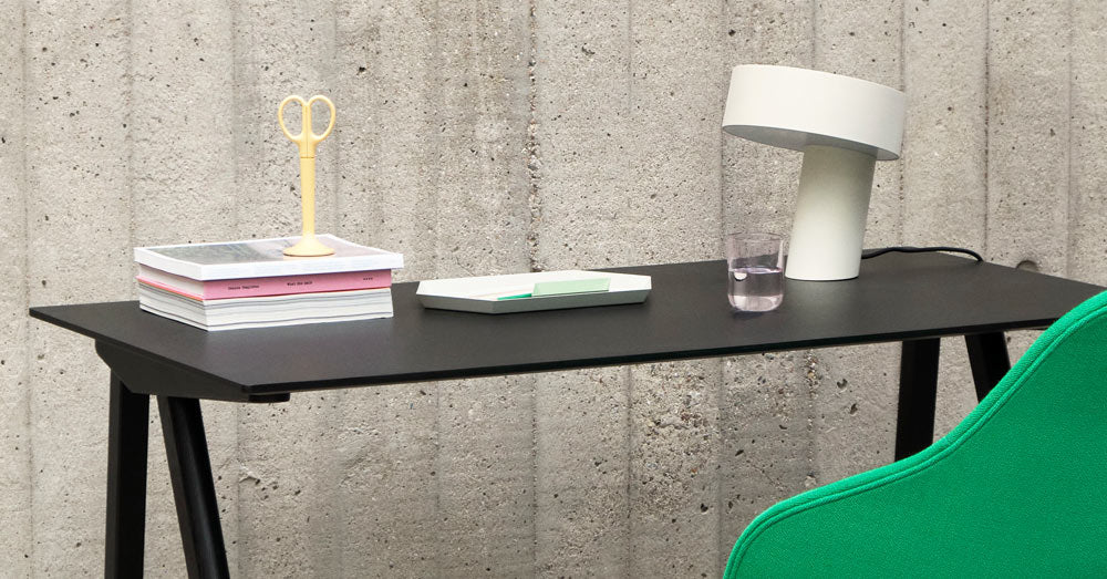 Une lampe Slant blanche de HAY, placée sur un bureau Copenhague 90 noir de HAY, à côté d'une pile de livres, de ciseaux jaunes et d'un verre d'eau.