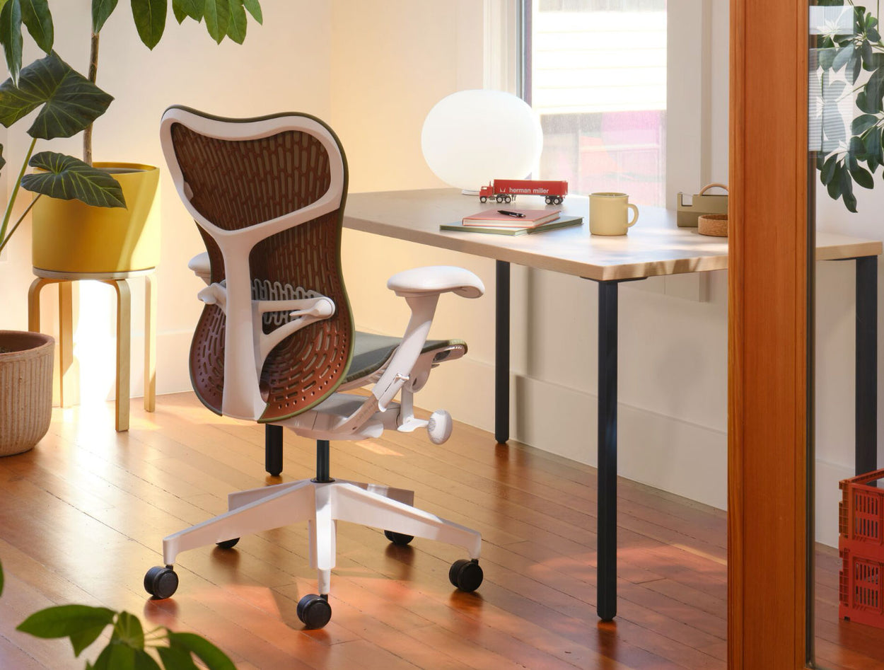 Une chaise de bureau Mirra 2 vert olive et blanche avec un dossier marron, placée devant un bureau Herman Miller OE1. Le tout dans un environnement de bureau à domicile, avec un sol en bois massif, des murs blancs et un éclairage chaleureux.