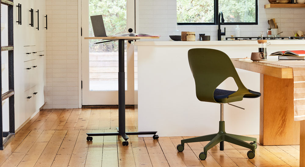 Une table Passport noire et chêne de Herman Miller placée près d'un îlot de cuisine blanc, avec un ordinateur portable et un livre. L'image montre également une chaise Zeph verte et noire d'Herman Miller placée près d'une table de salle à manger en bois.