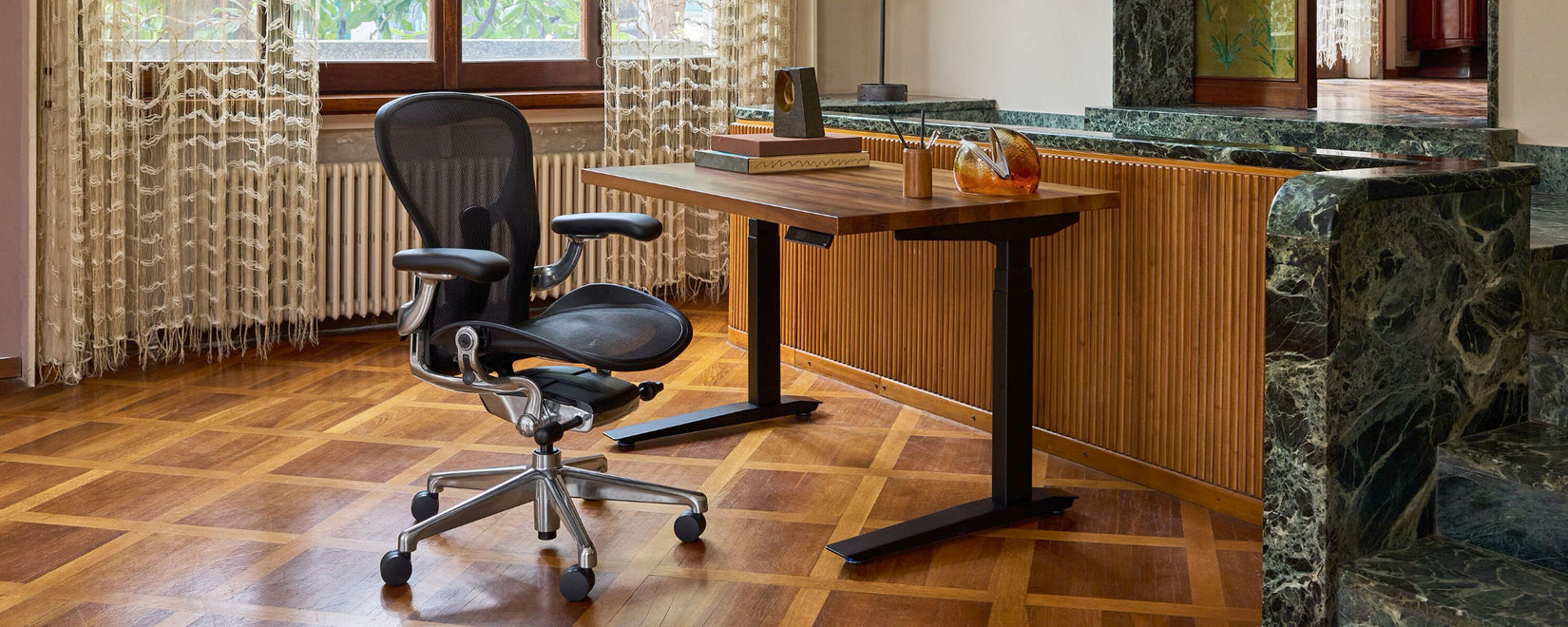 Un siège Aeron Herman Miller en aluminium poli noir onyx à côté dʼun bureau assis-debout Jarvis avec plateau en noyer dans un bureau à domicile de style méditerranéen avec boiseries et marbre noir émeraude.