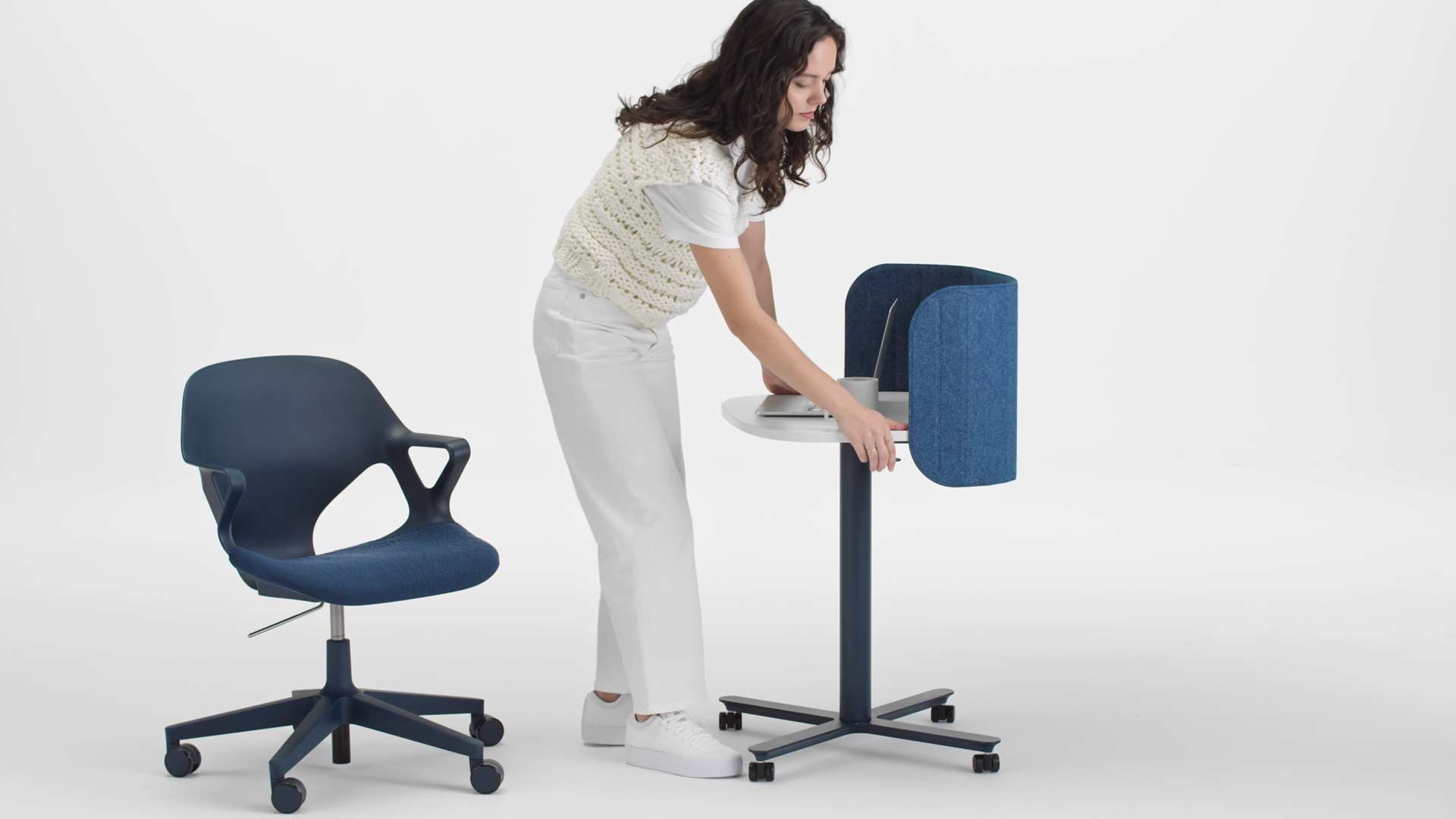 Vidéo d'une femme assise dans un fauteuil de bureau Zeph bleu qui se lève et élève la hauteur d'une table de travail Passport bleue et blanche de Herman Miller, en utilisant le levier situé sur le côté.