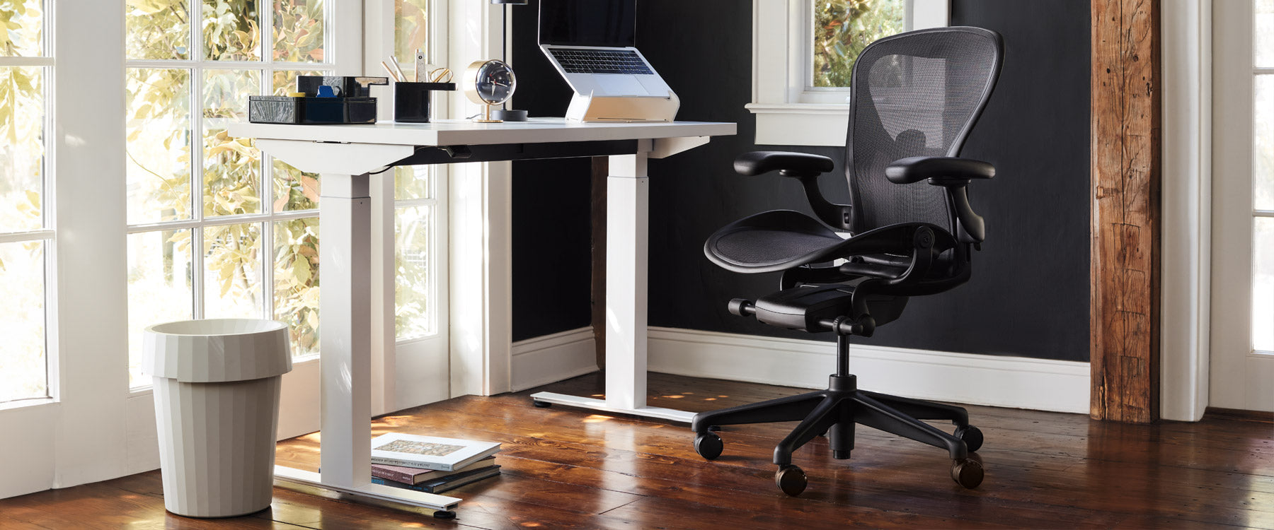 Scène de bureau à domicile avec un bureau assis-debout Nevi blanc, une chaise de bureau Aeron en noir onyx, un support pour ordinateur portable Oripura et une poubelle Hay Shade en blanc cassé