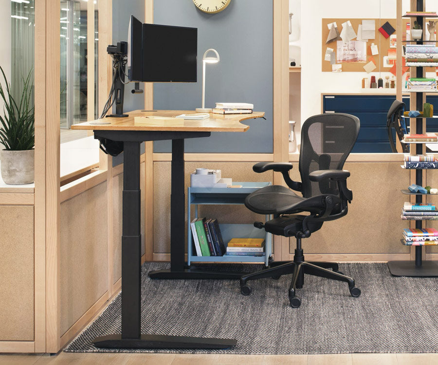 Home office setting showing Jarvis sit-stand desk in a standing position with black legs and contoured bamboo top alongside an Aeron office chair in graphite in background