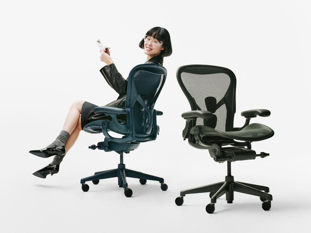 Dans un studio blanc, une femme était assise sur un fauteuil de bureau ergonomique Herman Miller Aeron bleu « Nighfall », à côté d'un autre Aeron vert « Jasper ».
