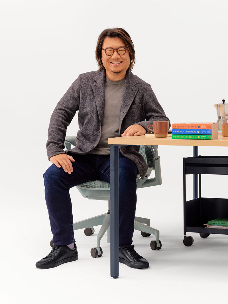 Photo joyeuse de Kevin Kwan assis sur un fauteuil de bureau Cosm gris glacier, appuyé contre un bureau OE1 à hauteur fixe avec des pieds couleur nuit et un plateau en frêne clair