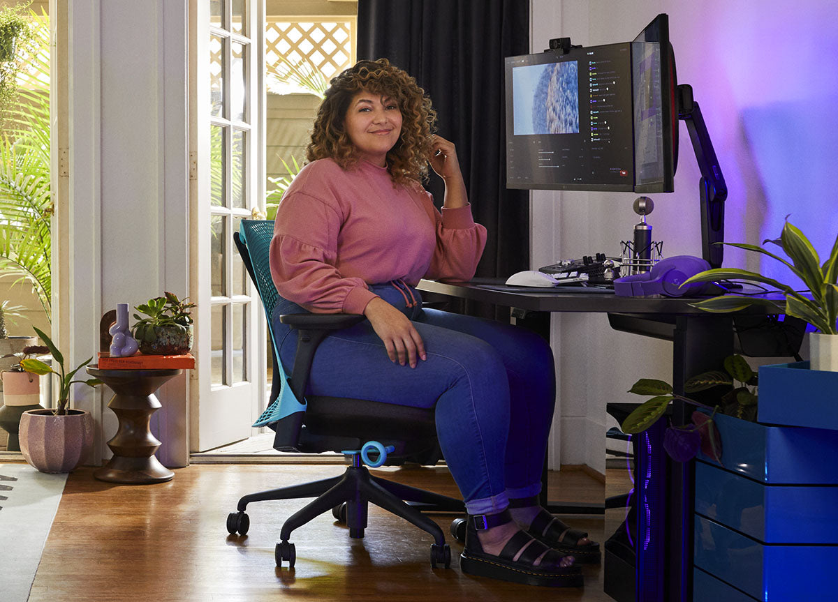 Une femme assise dans un fauteuil de jeu Sayl bleu et noir de Herman Miller, placé derrière un bureau Nevi noir avec un bras de moniteur et un écran Ollin noir. Le tout dans un espace intérieur avec des sols en bois, des murs blancs et des plantes d'intérieur.
