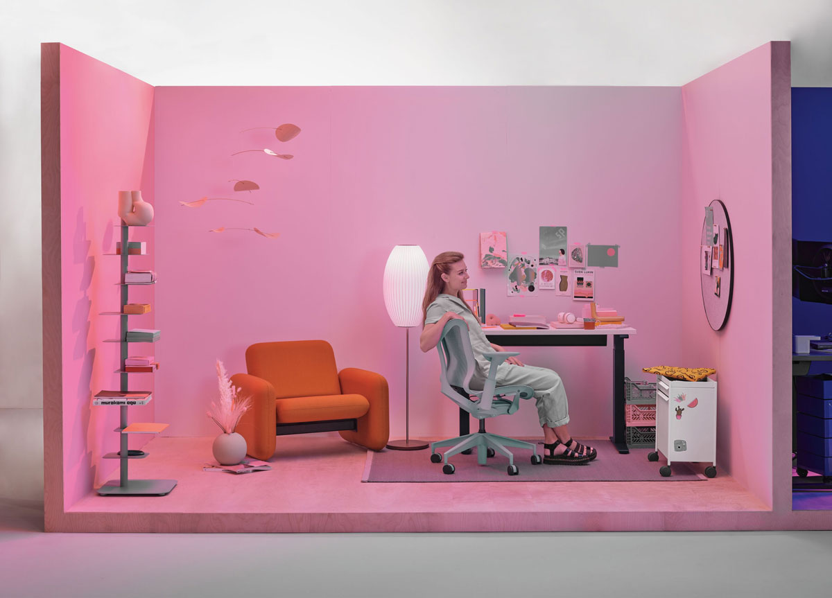 Bureau à domicile avec une femme assise sur une chaise de bureau Cosm à dossier bas en gris glacier, avec un bureau assis-debout compact et une chaise Wilkes en arrière-plan.