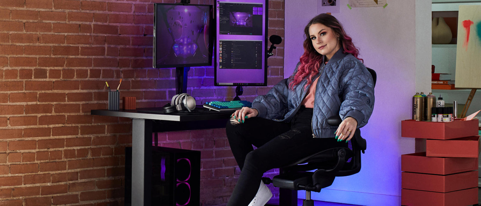 Photo de la joueuse Electra face à la caméra, assise sur une chaise de jeu Aeron noir onyx devant un bureau de jeu Nevi noir avec un bras support pour écran Ollin