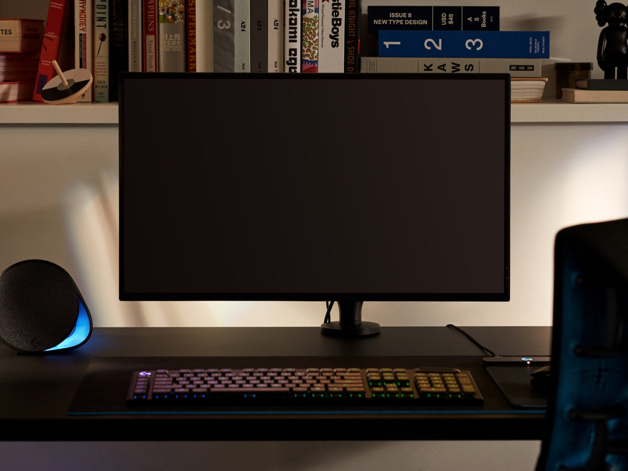 Vue de face d'un bras de moniteur Ollin noir tenant un écran d'ordinateur et fixé à un bureau de jeu Nevi noir de Herman Miller. Installé dans une salle de jeux ou un bureau à domicile, avec une étagère, un clavier et une souris.