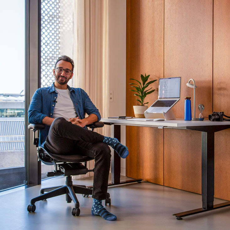 Photo d'Ali Abdaal, podcasteur, assis sur une chaise de bureau Herman Miller Aeron en graphite devant un bureau assis-debout Nevi avec des pieds noirs et un plateau blanc, un ordinateur portable posé sur un support pour ordinateur portable Oripura