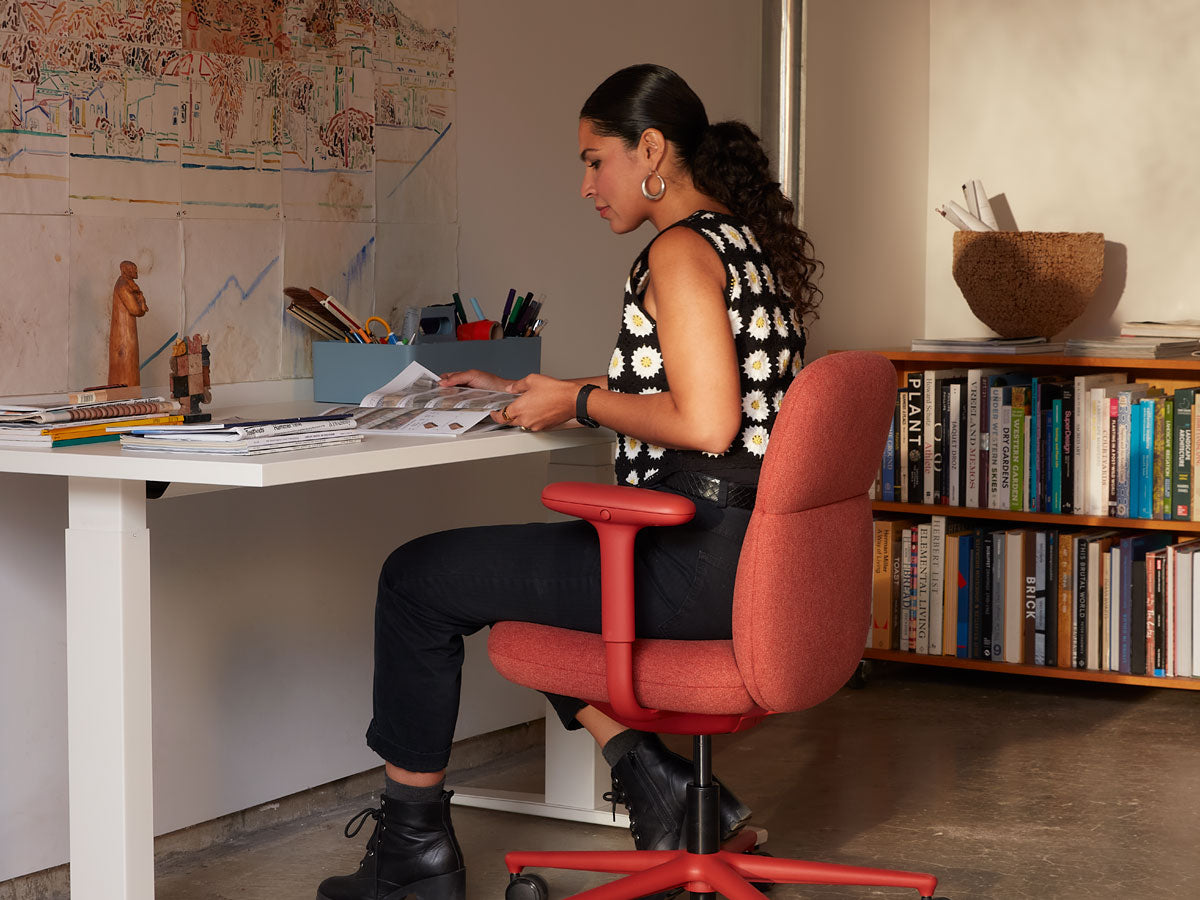 Anooshey Rahim assis, travaillant sur un fauteuil de bureau Asari à dossier moyen, couleur rouge canyon, devant un bureau assis-debout Nevi