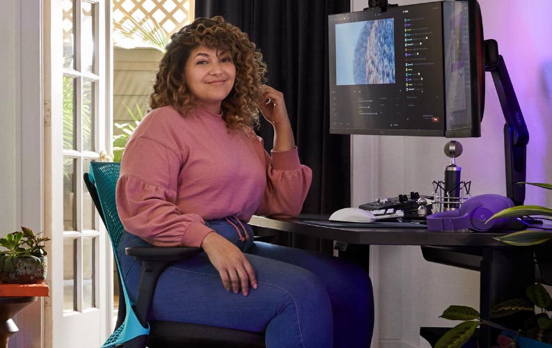 Le streamer professionnel et doubleur Friskk assis sur une chaise de jeu Herman Miller Sayl en bleu océan et noir, devant un bureau de jeu assis-debout Nevi noir avec un écran sur un bras de moniteur Oliin.