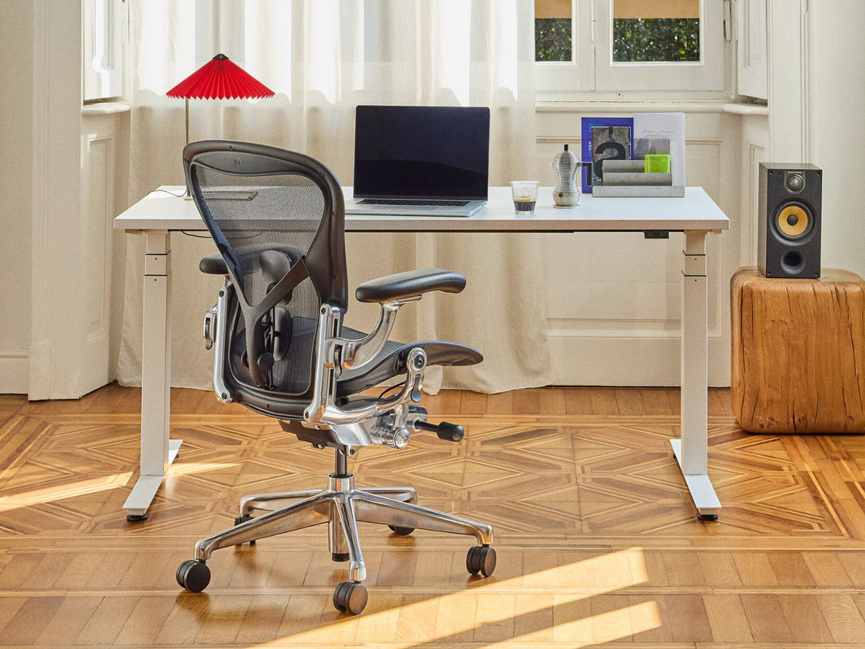 Un fauteuil de bureau ergonomique Herman Miller Aeron noir et chromé, intégré à un bureau à domicile lumineux comprenant un bureau assis-debout Nevi blanc.