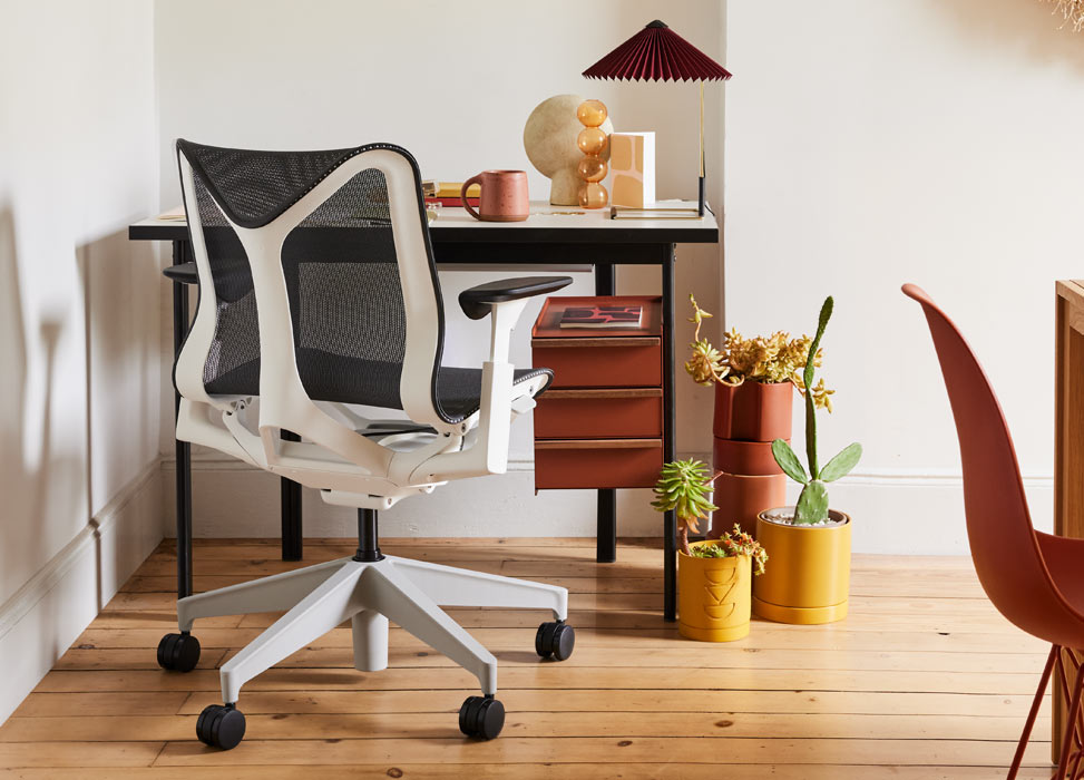 Un fauteuil de bureau Cosm bleu et blanc d'Herman Miller, placé devant un bureau Mode noir et terre cuite d'Herman Miller. Dans un bureau à domicile au sol en bois et aux murs blancs, accessoirisé d'une lampe de table Matin et de plantes d'intérieur.