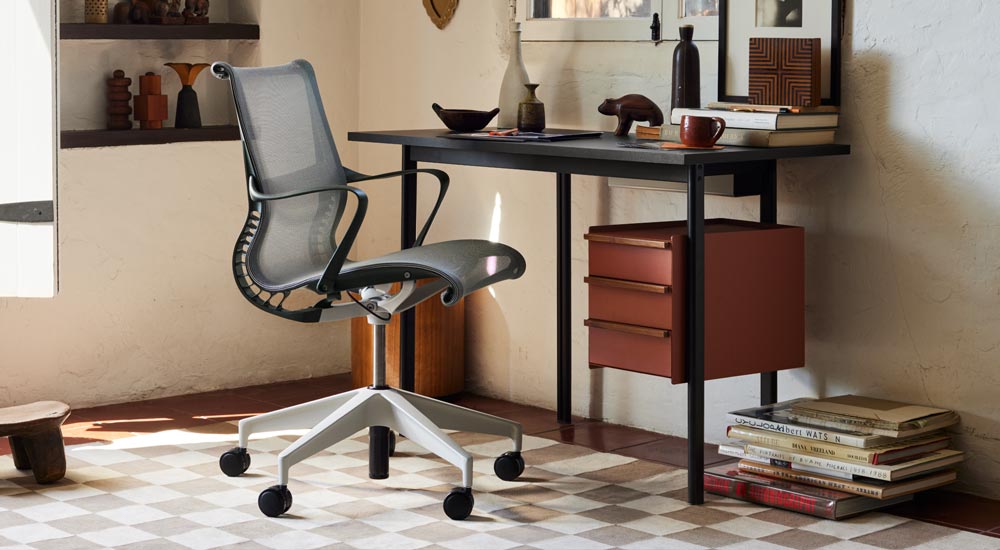 Un bureau Mode noir d'Herman Miller, avec des rangements en terre cuite et des poignées en noyer, est placé devant une chaise de bureau Setu grise d'Herman Miller. Un bureau bien meublé avec des étagères, des livres et des accessoires décoratifs.
