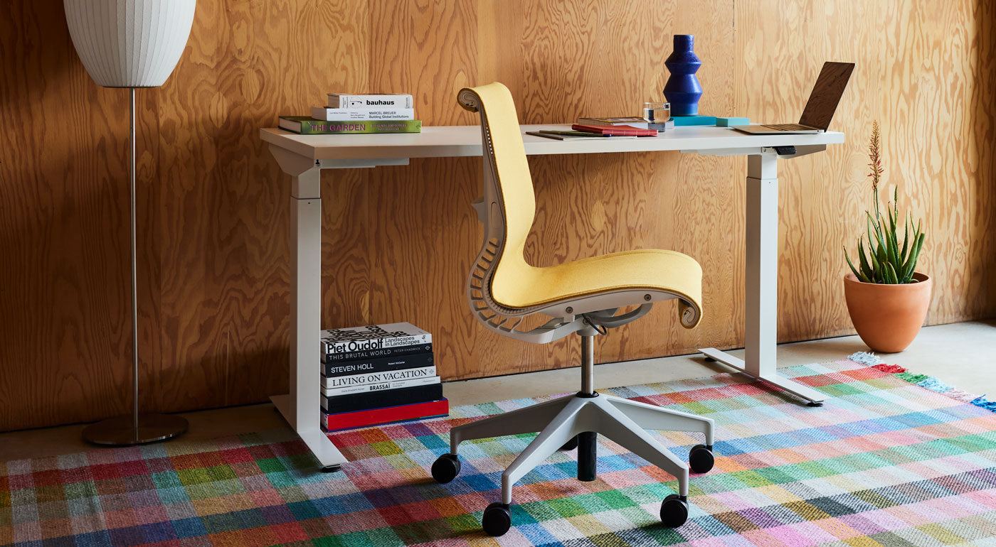 Un siège de bureau Setu jaune d'Herman Miller, revêtu de tissu Mimosa Meld, placé devant un bureau debout Nevi blanc sur un tapis à carreaux coloré.