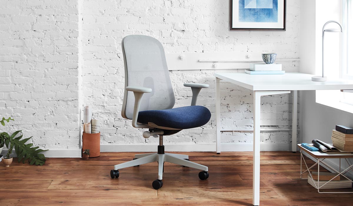 Un siège de bureau Lino gris et bleu de Herman Miller est placé devant un bureau blanc. L'espace intérieur se caractérise par un mur de briques blanches, un sol en bois et une lampe de table Marselis blanche posée sur le bureau.