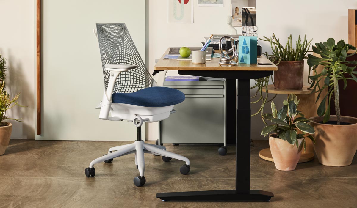 Un fauteuil de bureau Sayl blanc et bleu d'Herman Miller, placé à côté d'un bureau debout Jarvis avec des pieds noirs et un plan de travail en bambou. Dans un environnement de bureau à domicile, avec un chariot OE1 bleu et une sélection de plantes d'intérieur dans des pots en terre cuite.