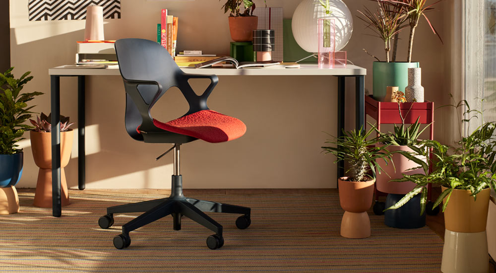Un siège de bureau Zeph noir avec une assise rouge est placé devant un bureau Herman Miller OE1. Dans un environnement de bureau à domicile, avec un chariot OE1 rouge et une sélection de plantes d'intérieur dans des pots colorés.