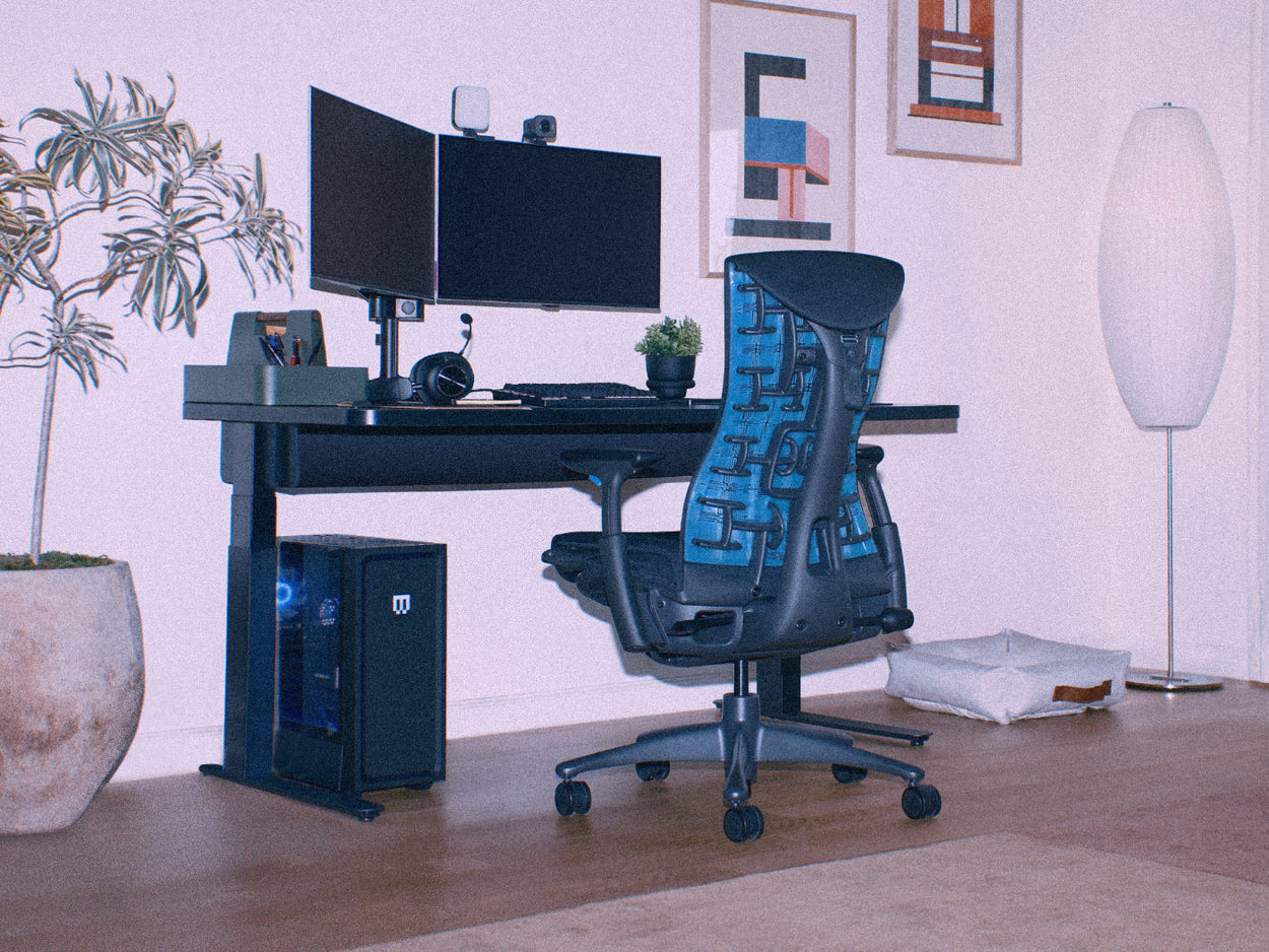Une image un peu granuleuse d'un fauteuil gaming Herman Miller Embody noir et bleu cyan, faisant partie d'une installation de gaming sur PC à domicile comprenant un bureau gaming Nevi réglable en hauteur.