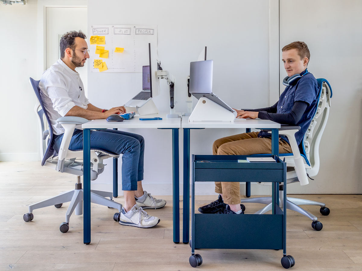 Photo de deux hommes assis à un bureau l'un en face de l'autre, montrant que lorsque vous êtes assis correctement, vos coudes sont au niveau du dessus du bureau. Caractéristiques : bureau OE1 avec pieds bleus et plateau blanc, chaise Mirra 2 en blanc minéral et bleu et chaise Embody en blanc et bleu