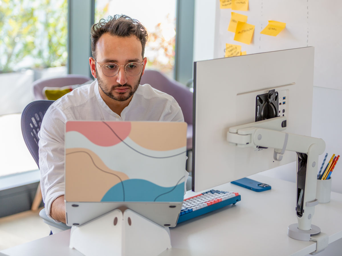 Photo d'un homme assis à un bureau avec un écran devant lui sur un bras support d'écran Ollin et un ordinateur portable sur un support Oripura, montrant que les yeux doivent être directement en face de l'écran, aux deux tiers de la hauteur de celui-ci.