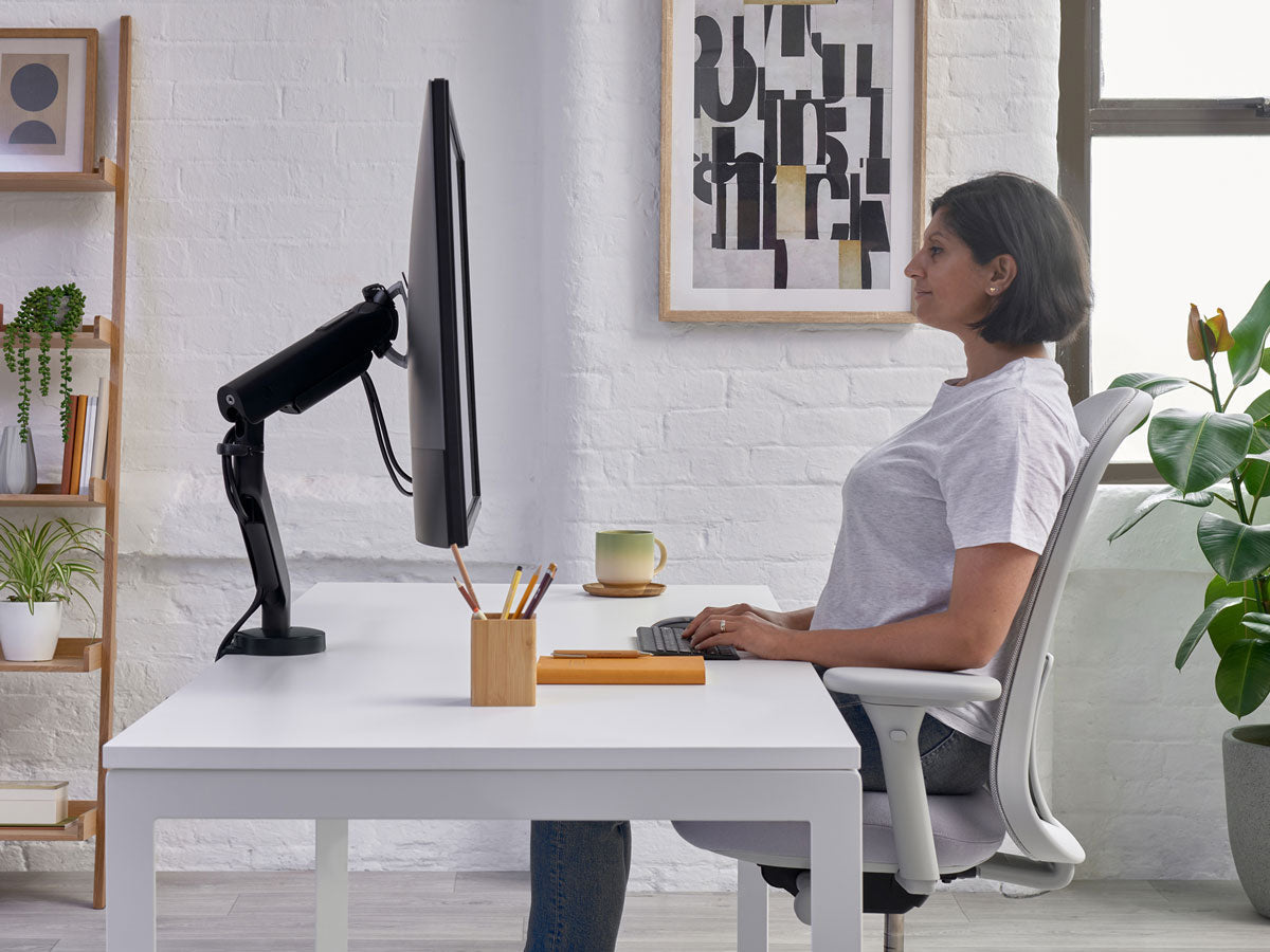 Femme assise à un bureau sur une chaise de bureau Lino en blanc minéral avec assise grise, montrant que ses poignets sont au niveau du bureau, ce qui correspond à une position assise correcte