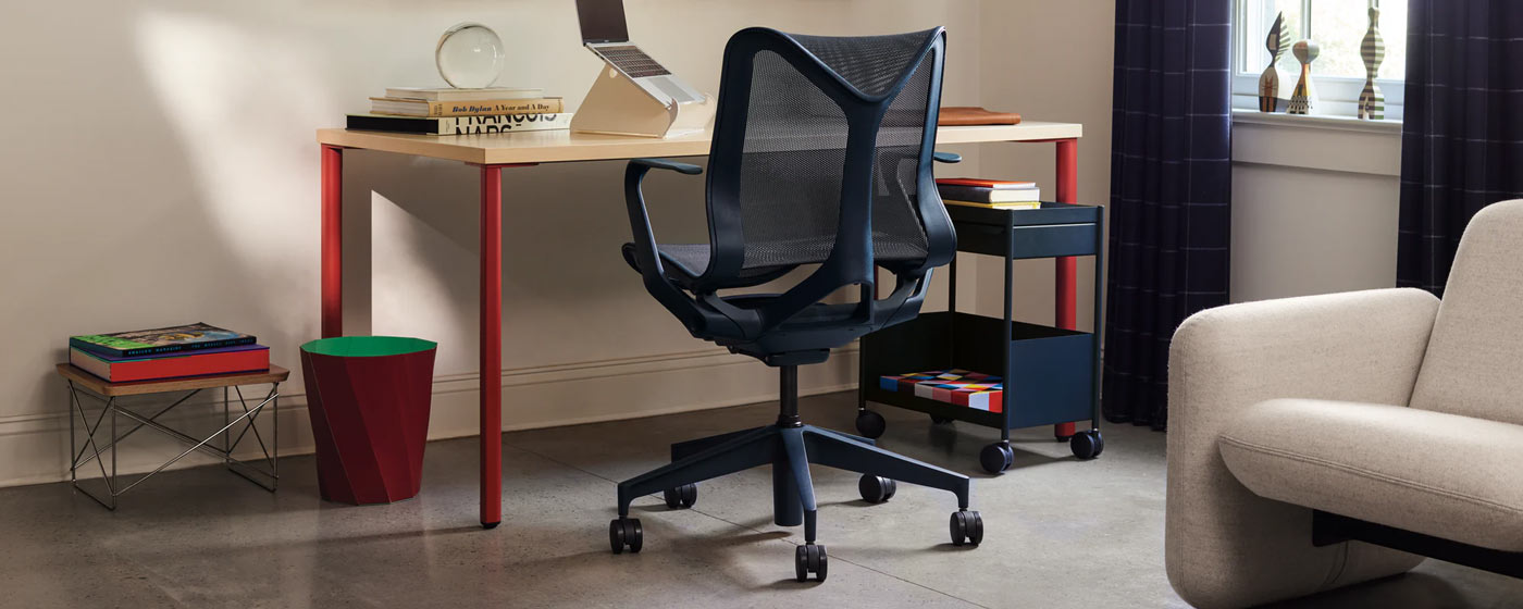 Une chaise de bureau Cosm bleue de Herman Miller, placée devant un bureau OE1 rouge et gris cendré de Herman Miller. Installée dans un bureau à domicile, elle est accessoirisée d'un chariot OE1 bleu, d'un support pour ordinateur portable Oripura blanc et d'une corbeille à papier HAY Paper rouge.