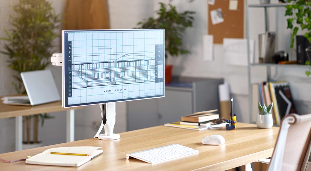 Un bras de moniteur Ollin blanc de CBS, monté sur un bureau en bois supportant un écran d'ordinateur, dans un environnement de bureau.
