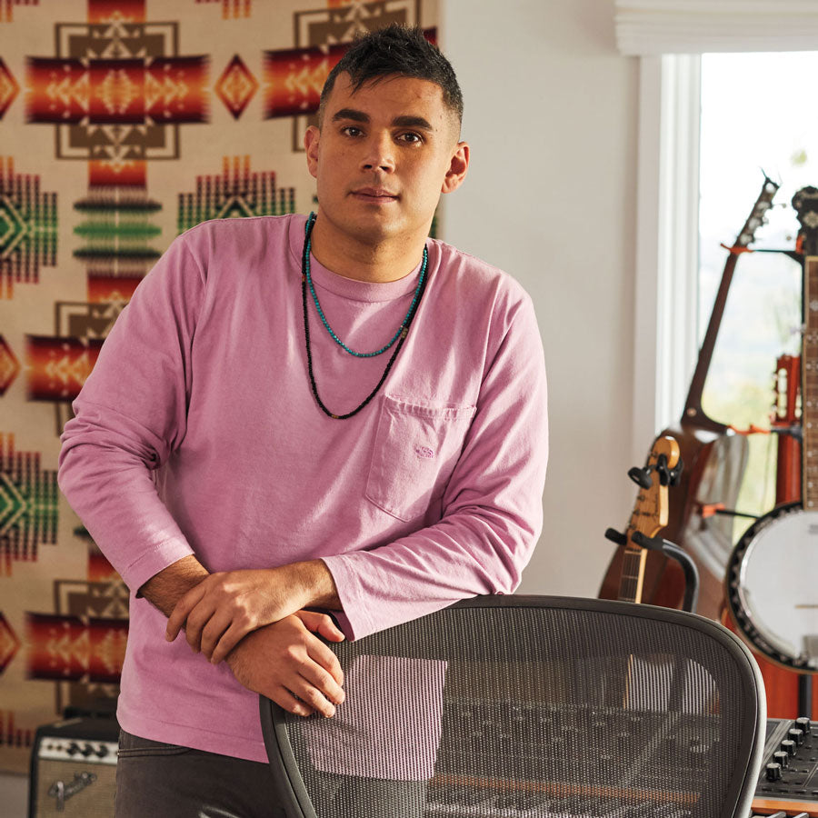 Photo de Rostam, musicien, producteur et compositeur, debout appuyé contre une chaise de bureau Aeron en graphite, avec des instruments de musique en arrière-plan