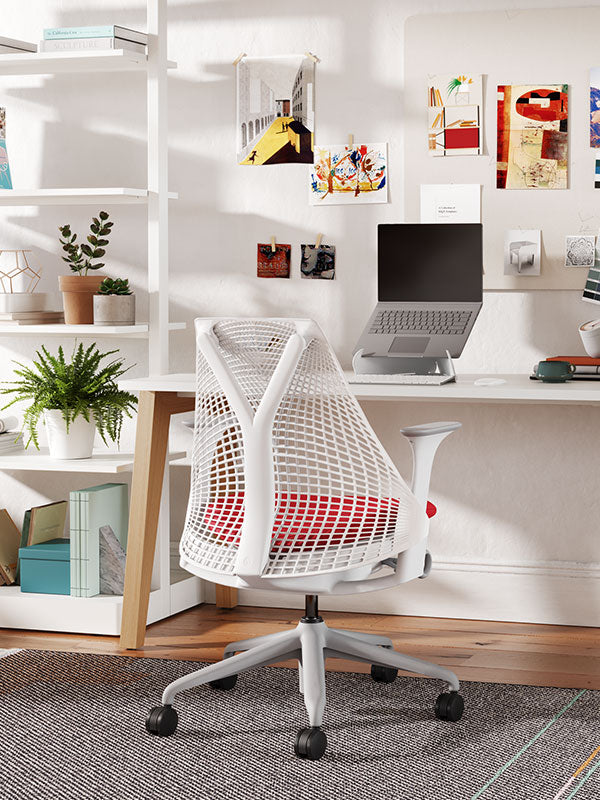 Chaise de bureau Sayl avec structure blanche, base grise et assise rembourrée rouge dans un bureau à domicile, avec étagères, plantes et ordinateur portable sur un support Orpura blanc.