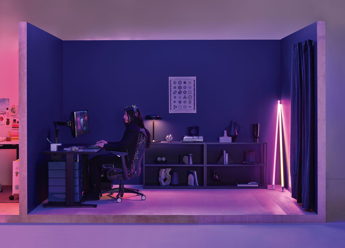Bureau dédié au gaming avec une femme assise sur une chaise de gaming Logitech Embody noire, devant un bureau gaming Nevi noir et un tube néon Hay dans le coin.