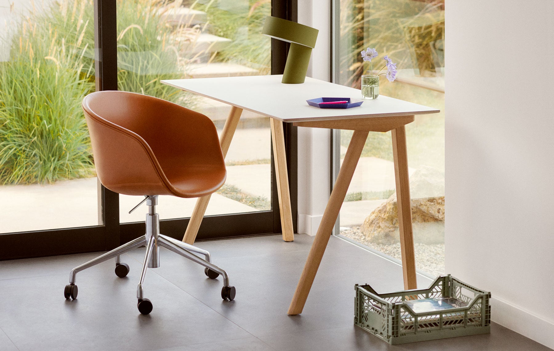 Photo simple dans un cadre domestique avec vue sur le jardin, montrant un bureau Hay Copenhague 90 en chêne avec plateau blanc, une lampe Hay Slant vert kaki sur le bureau et une petite caisse de rangement Hay au sol