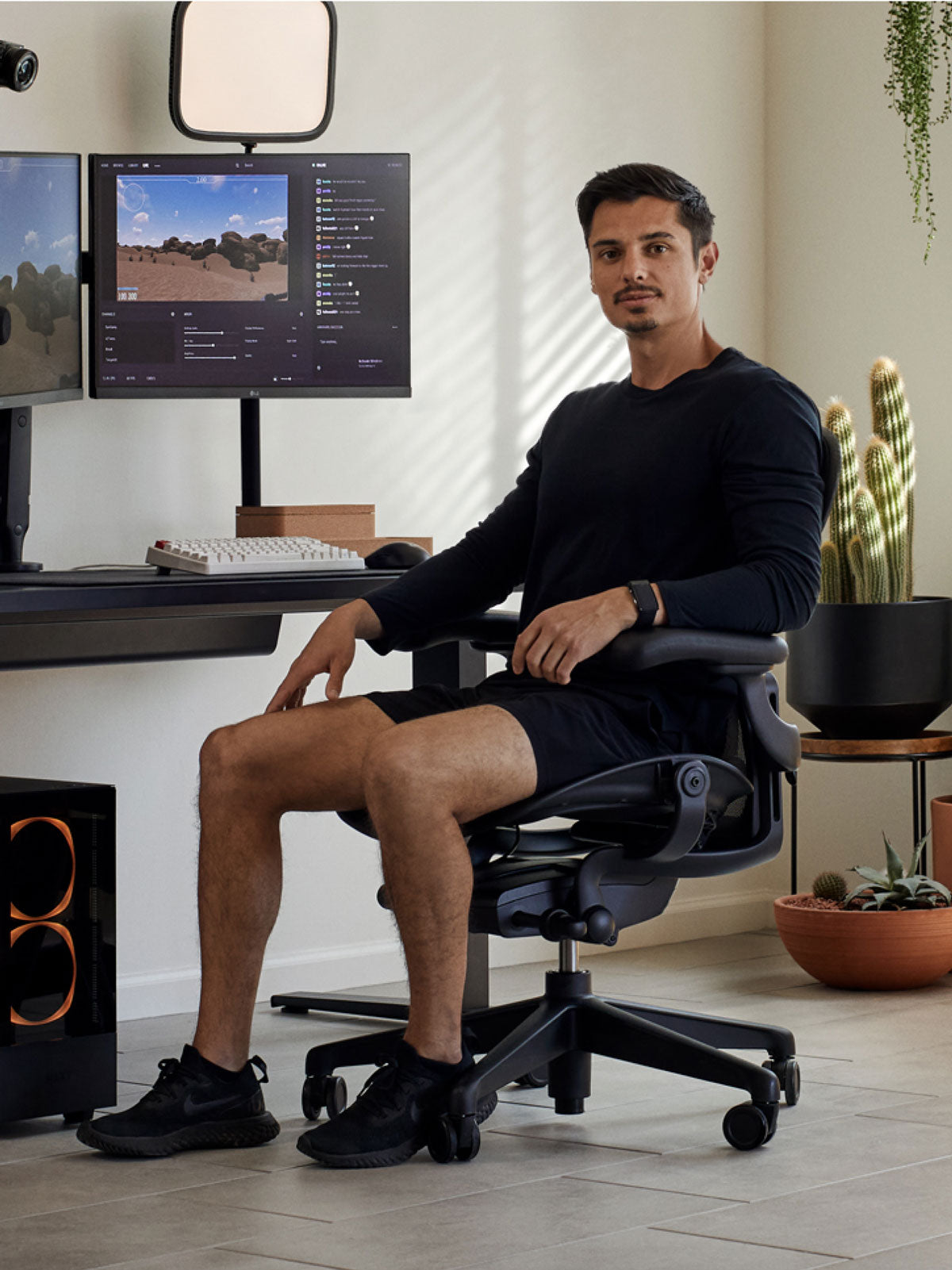 Un homme assis dans un fauteuil de jeu Aeron noir de Herman Miller, placé derrière un bureau Nevi noir avec un bras de moniteur et un écran Ollin noir. Le tout dans un espace intérieur avec des sols carrelés, des murs blancs et des plantes d'intérieur dans des pots en terre cuite.