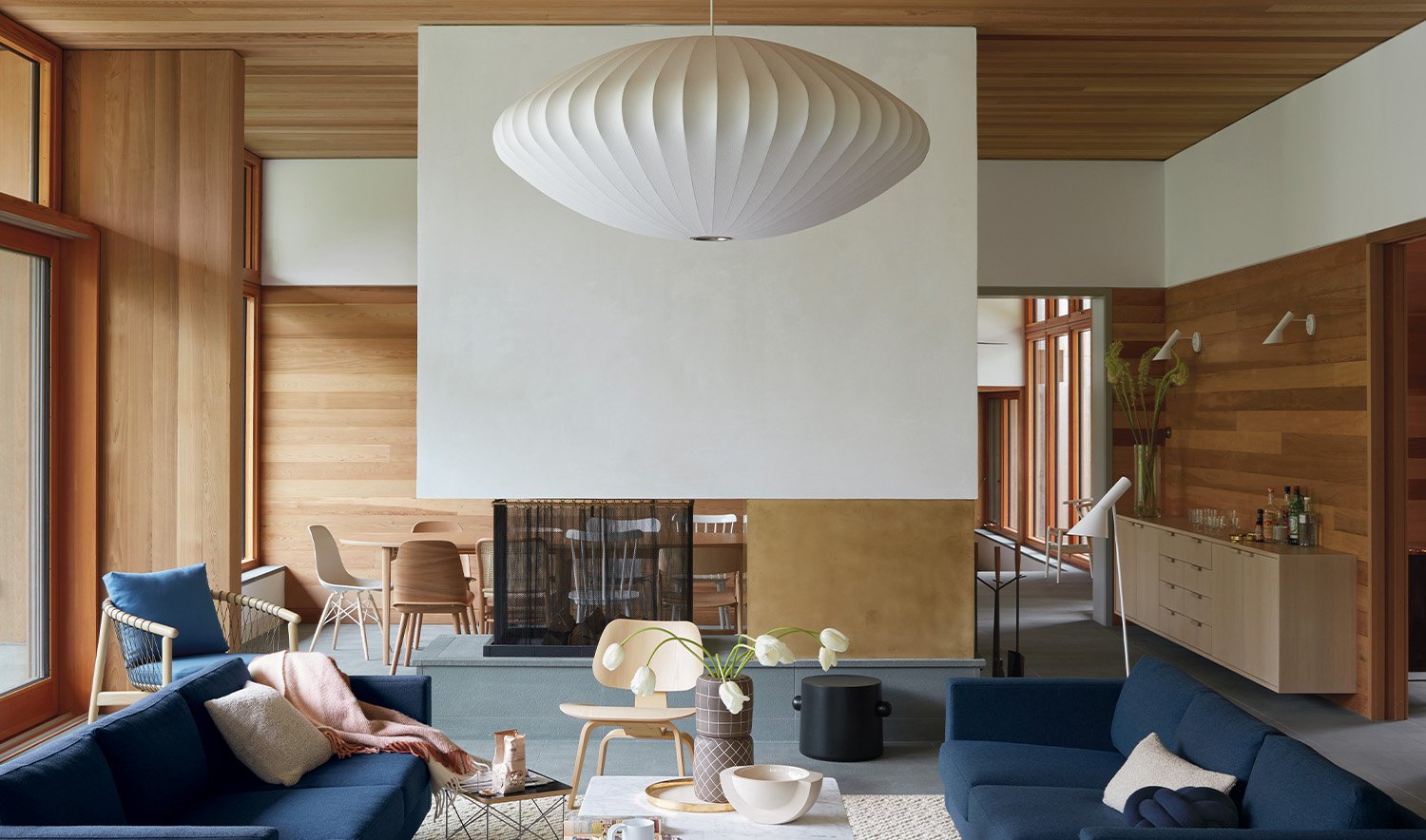 Un salon moderne avec une cheminée contemporaine, avec une grande suspension Nelson bubble blanche de Herman Miller. La pièce est remplie de meubles, dont deux canapés bleus, des chaises d'appoint, un buffet et une table basse.