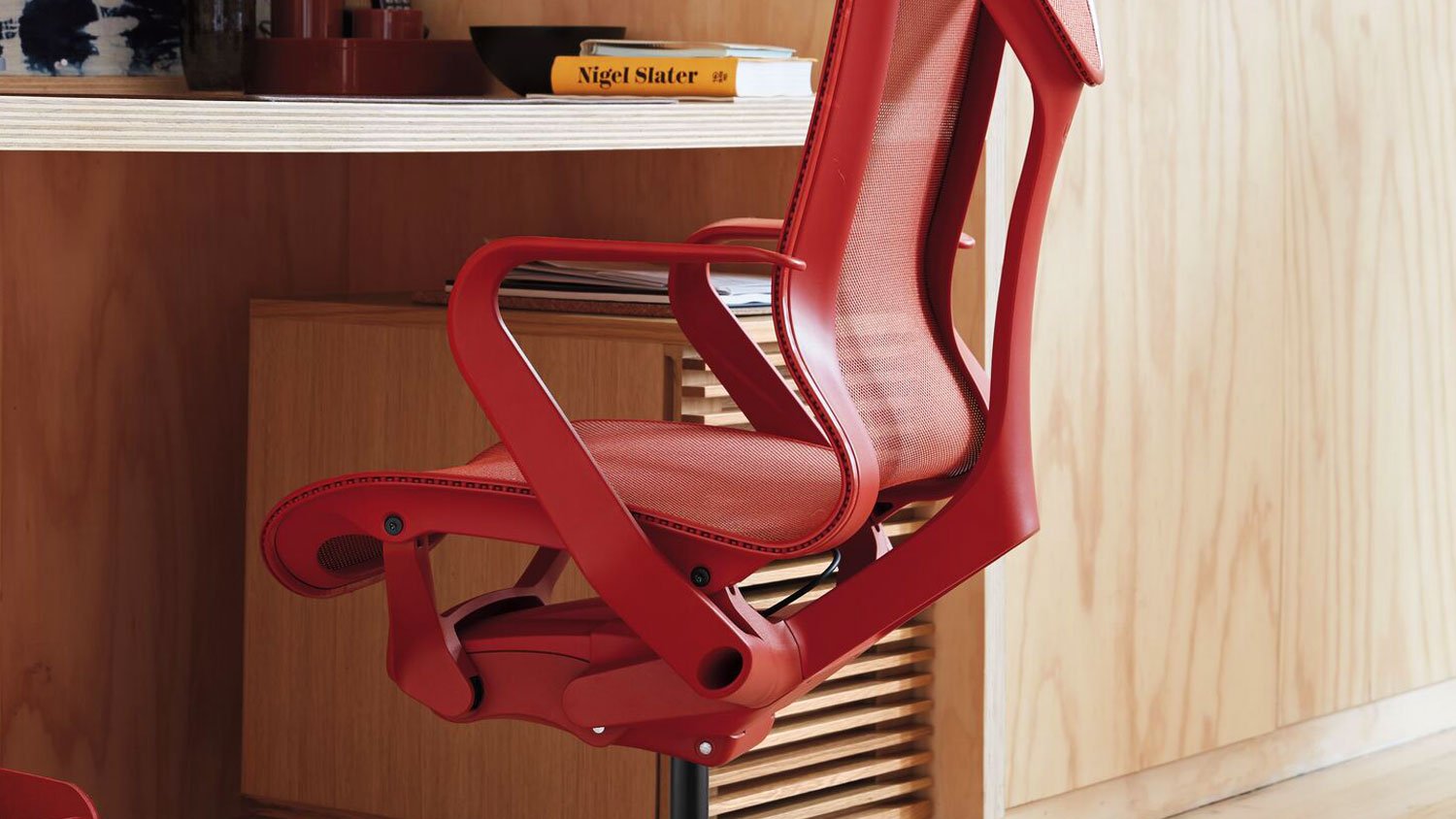 Une chaise de bureau Cosm rouge de Herman Miller est placée sous un bureau. Elle est placée dans un environnement intérieur avec un sol et des murs en bois dur, l'accent étant mis sur la couleur vive de la chaise parmi les meubles qui l'entourent.