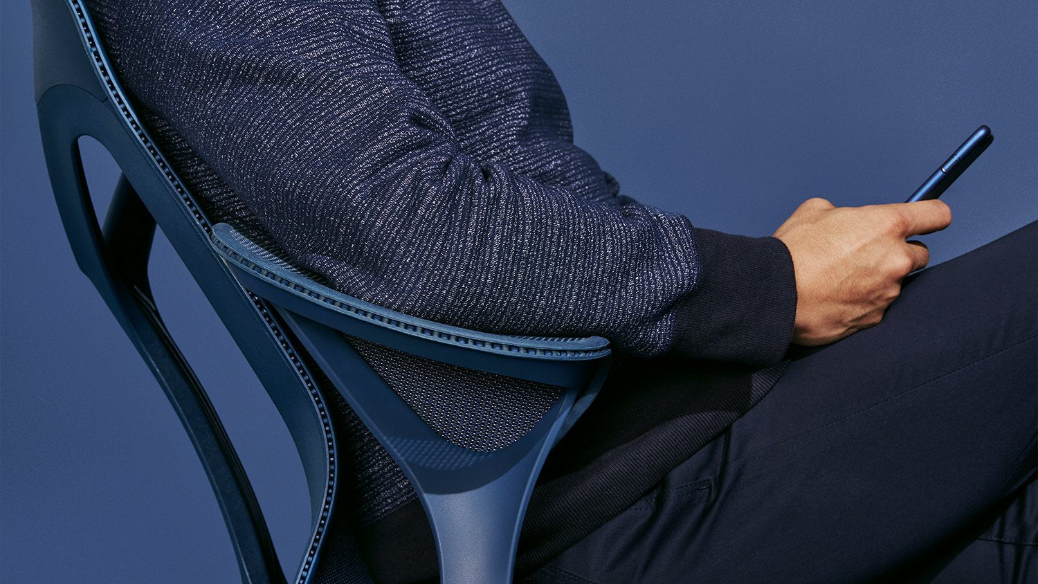 Une personne assise sur une chaise de bureau Cosm bleu nuit de Herman Miller. Elle tient un téléphone et est vêtue de vêtements bleu foncé devant un fond bleu foncé uni.