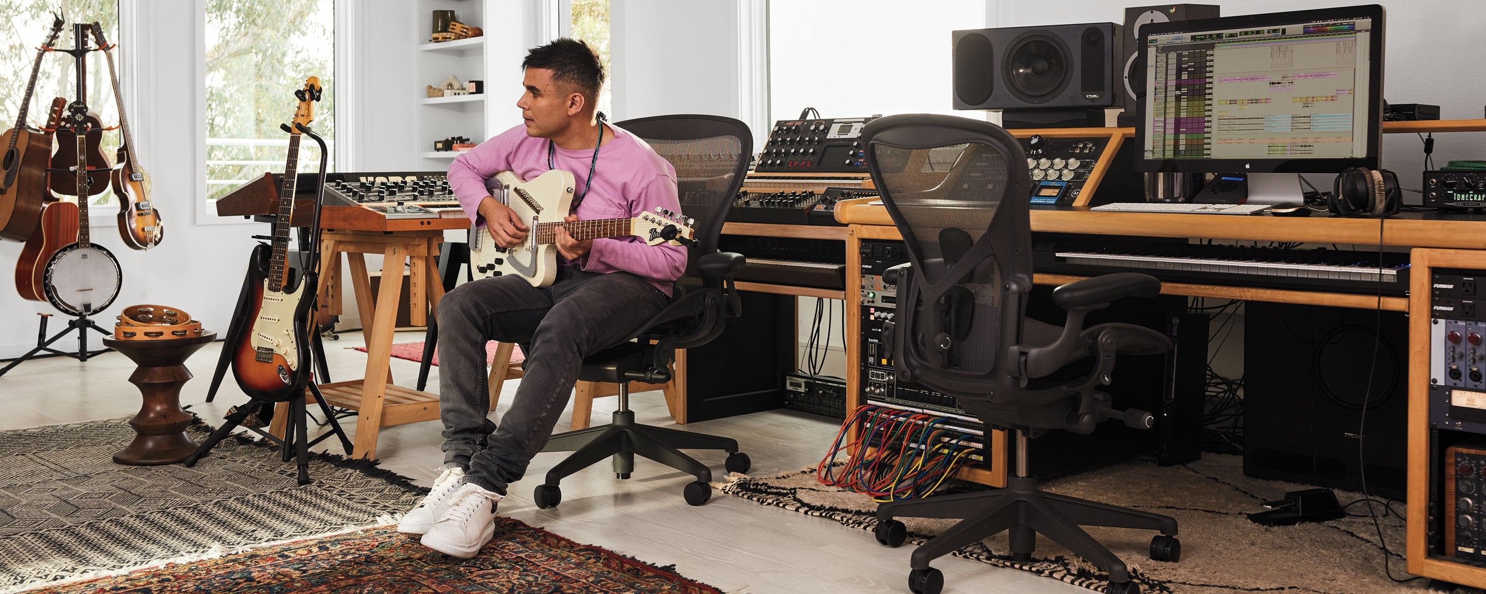 Photo de Rostam, musicien, producteur et compositeur, dans son studio de musique, jouant de la guitare, assis sur une chaise de bureau Aeron en graphite