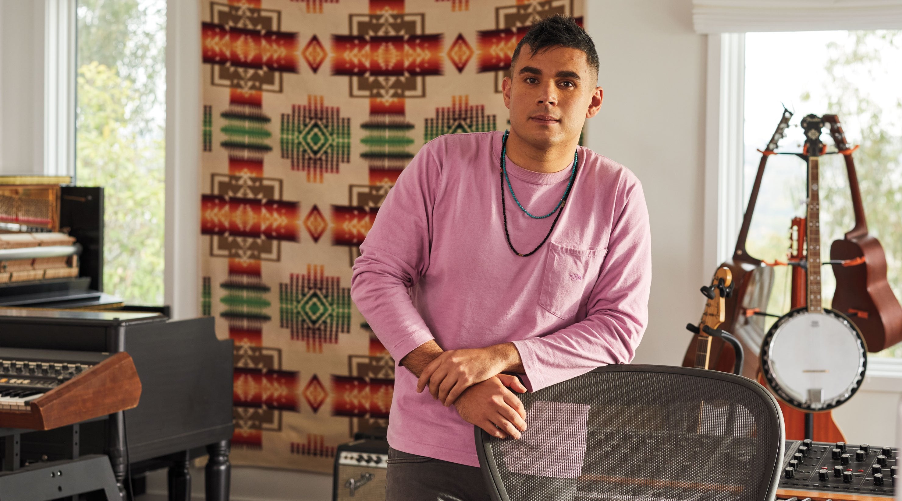 Photo de Rostam, musicien, producteur et compositeur, debout, appuyé sur une chaise de bureau Aeron en graphite, avec des instruments de musique en arrière-plan