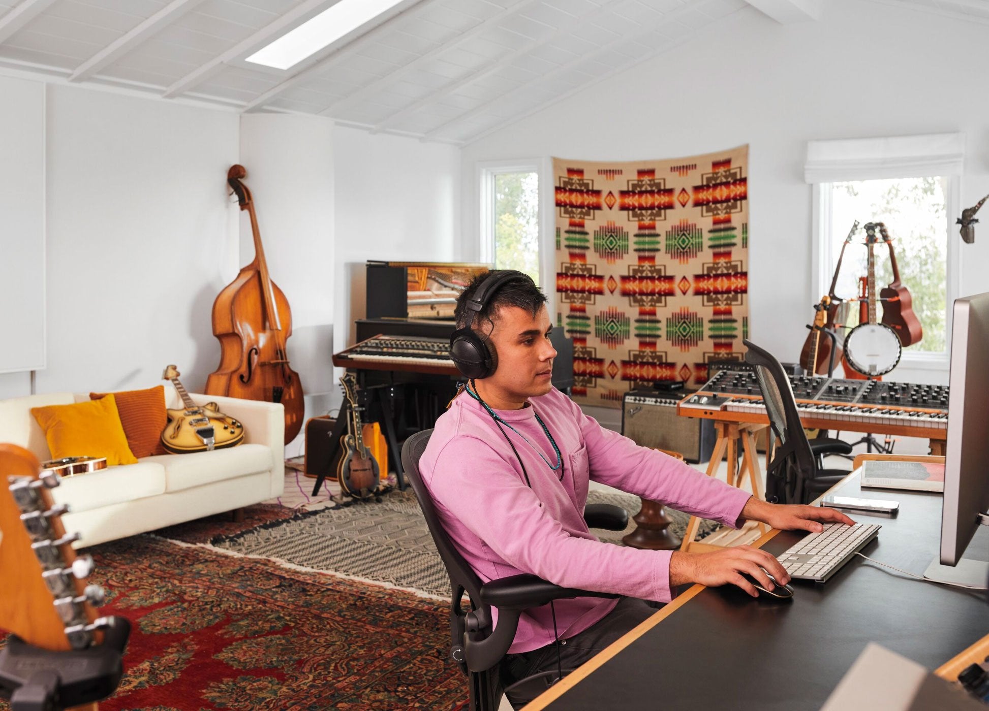 Photo de Rostam, musicien, producteur et compositeur, assis sur une chaise de bureau Aeron en graphite devant un ordinateur, avec un casque sur les oreilles et des instruments de musique en arrière-plan